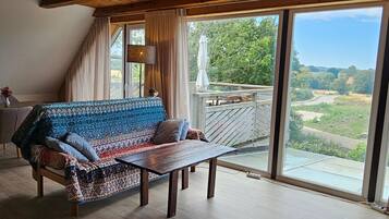 Ein Zimmer mit einer Sitzecke, einem Tisch und Blick auf eine Landschaft durch große Fenster.