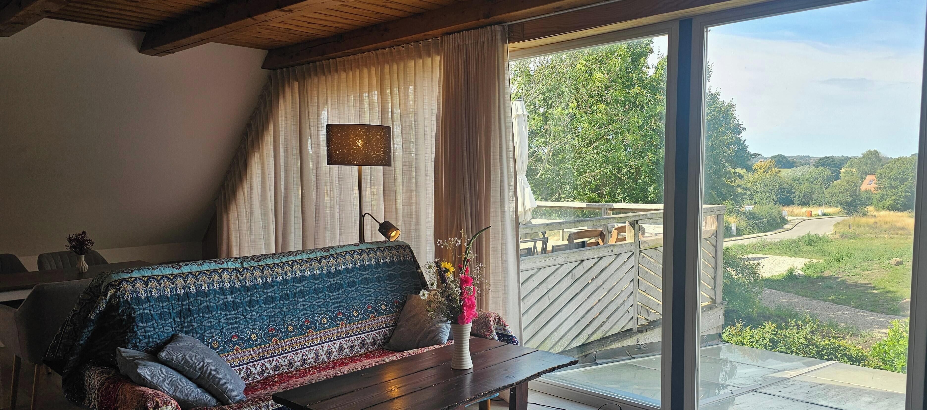 Ein Zimmer mit einer gemusterten Couch, einem Tisch mit Blumenvase und Blick auf eine grüne Landschaft durch ein großes Fenster.