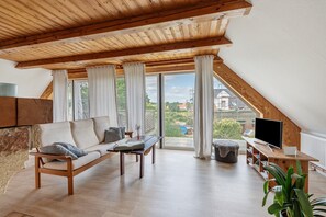 En rymlig loftvåning med ett träpanelat tak, en soffa, en tv och ett stort fönster med utsikt över omgivningen.