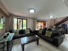 A living room with a sofa set, a coffee table, and a dining table with chairs. There is a staircase leading to an upper level.