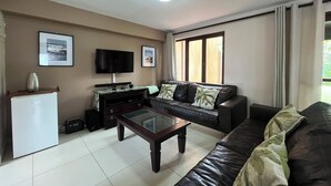 A living room with a television, a coffee table, a sofa, and a window with curtains. - San Lameer Villa 2802 (Southbroom)