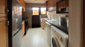 A compact kitchen with wooden cabinets, a stainless steel refrigerator, a washing machine, and a microwave. - San Lameer Villa 2802 (Southbroom)