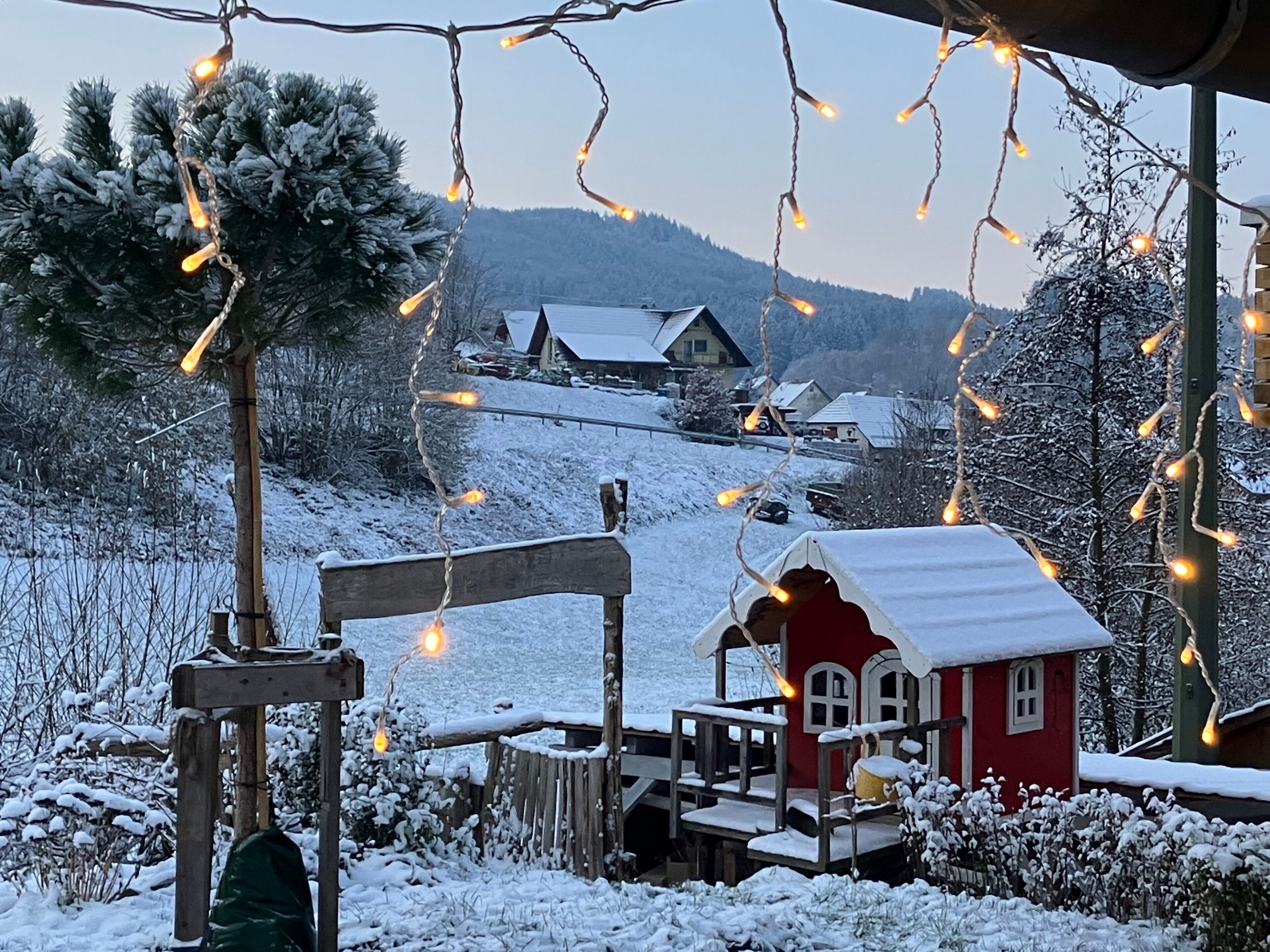 Eine verschneite Winterlandschaft mit einer hölzernen Bank, einem kleinen roten Haus und einem Baum.