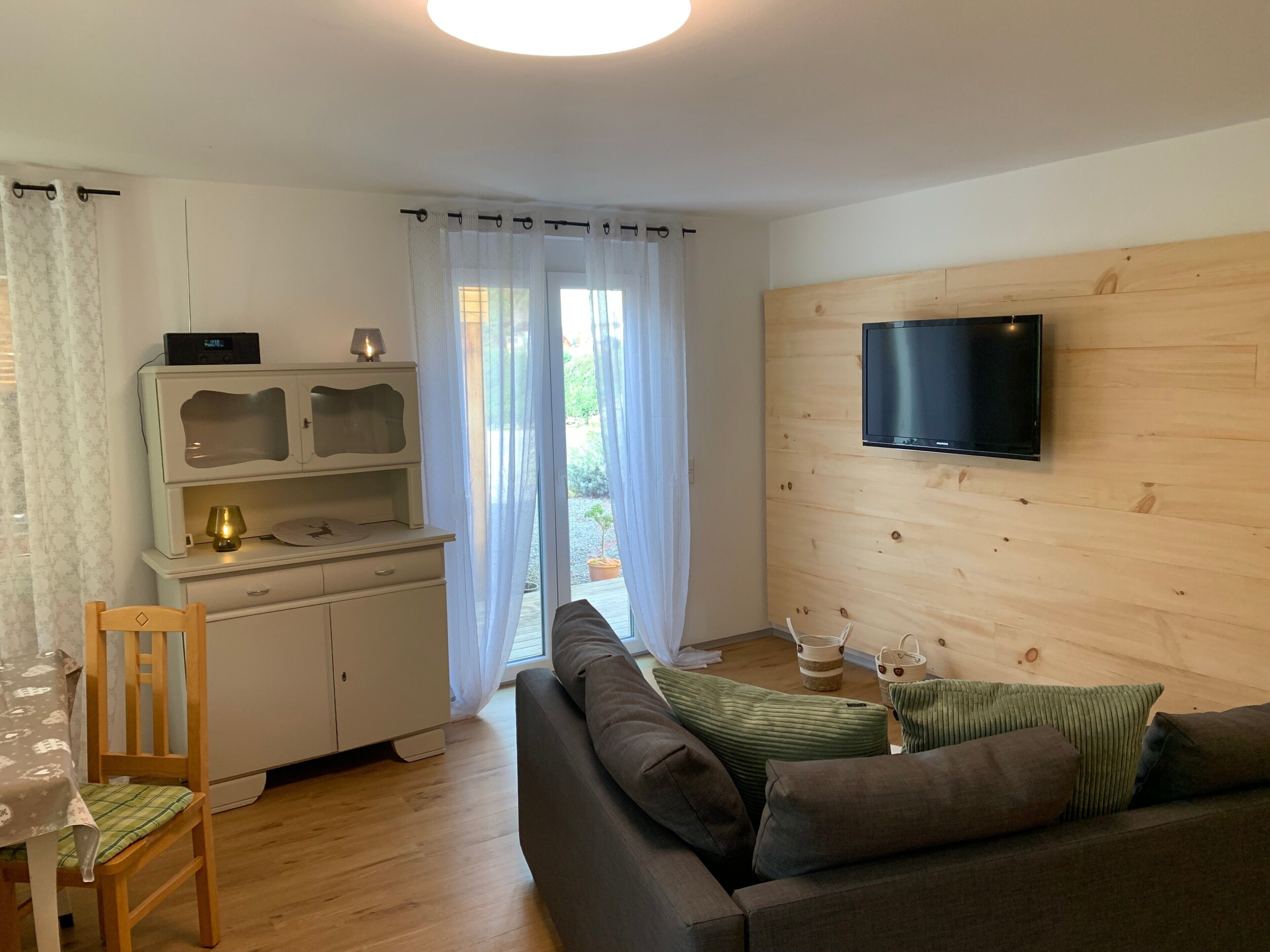 Un salon comprenant un canapé, un téléviseur fixé à un mur en bois, un coin repas avec une table et des chaises, et une porte-fenêtre coulissante donnant sur un espace extérieur.