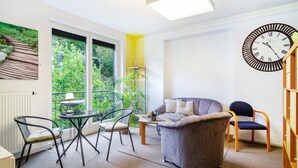 Ein modernes Wohnzimmer mit einer Couch, Sesseln, einem Tisch, einer Uhr und Blick durch große Fenster.