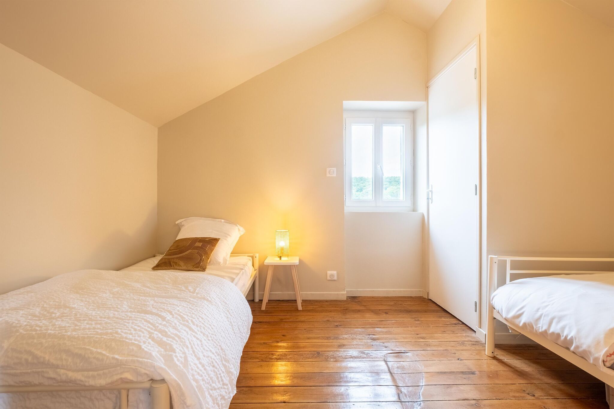 Ein Schlafzimmer mit zwei Betten, einem Nachttisch mit Lampe, Holzboden und einem Fenster, das natürliches Licht hereinlässt.