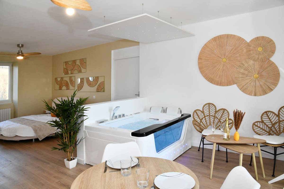 Une chambre d’hôtel moderne dotée d’une grande baignoire, d’un coin repas avec une table et des chaises, et d’une plante en pot.
