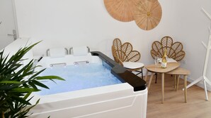 Een spa-ruimte met een jacuzzi, twee rieten stoelen, een klein tafeltje met een fles en glas, en een potplant.