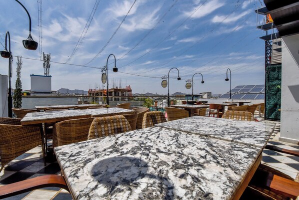 An outdoor dining area with marble tables and wicker chairs, overlooking a body of water and distant mountains.