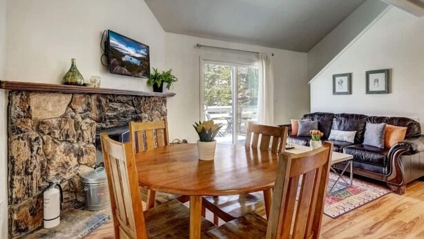 Un coin repas avec une cheminée en pierre, une table en bois et des chaises. On aperçoit un canapé en cuir noir et une fenêtre avec des rideaux au fond.