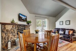 A dining area with a stone fireplace, wooden table, and chairs. There is a black leather sofa and a window with curtains in the background.