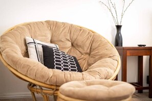 A rattan chair with cushions, a small wooden table, and a vase with branches.