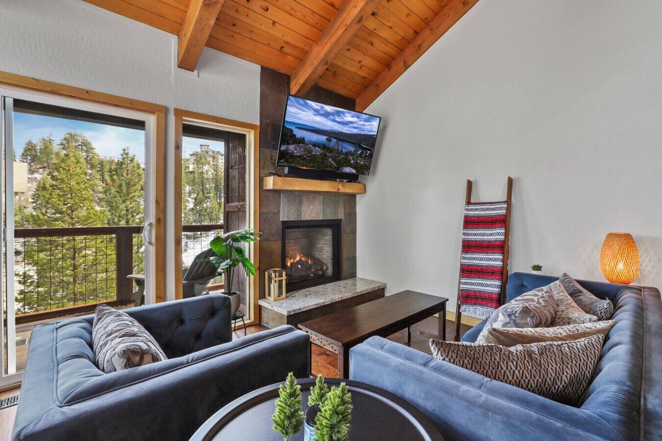 A modern living room with a fireplace, comfortable seating, a TV, and large windows overlooking a forested area.
