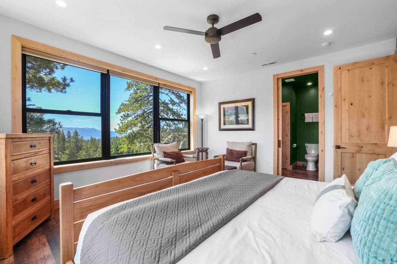 A bedroom with a large window, a bed, a dresser, a chair, and a ceiling fan.