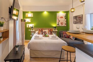 Une chambre d’hôtel moderne avec un grand lit, une petite table à manger et un banc. On y trouve des étagères avec des plantes et des livres, ainsi qu’un mur d’accent de couleur verte.