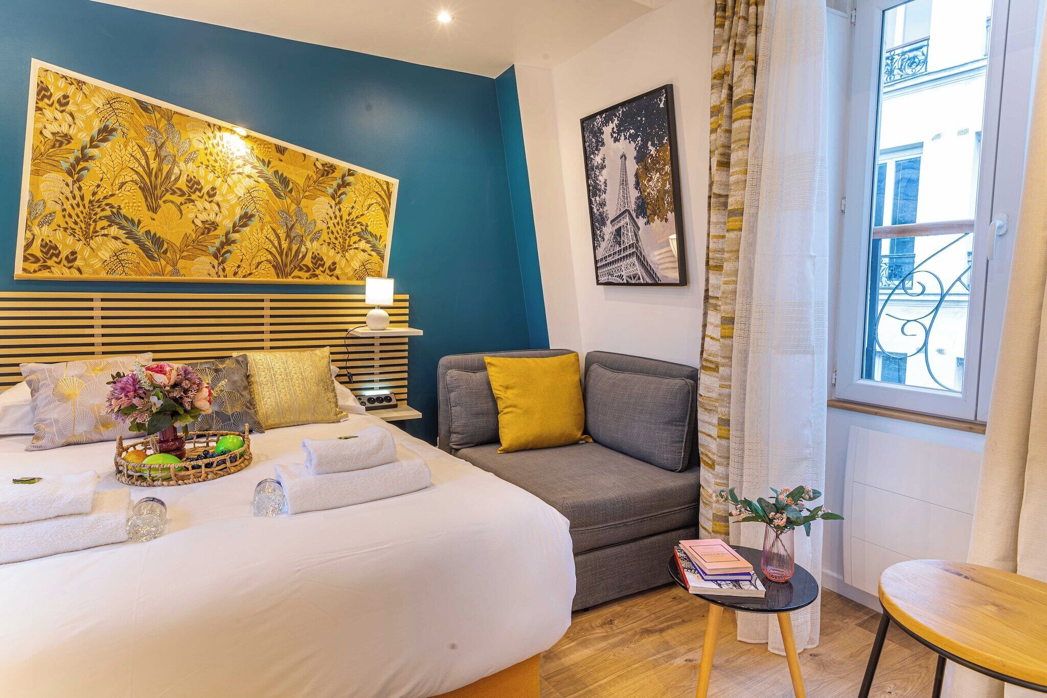 Una habitación de hotel con una cama, un sofá gris, una mesita con un jarrón y libros, una obra de arte floral grande en la pared y una fotografía enmarco de la Torre Eiffel.