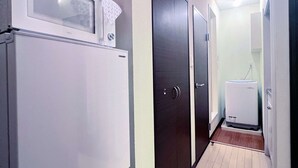 En smal hall med ett kylskåp och en liten vit apparat på vänster sida, en mörk dörr på höger sida och en ljusströmbrytare på väggen.