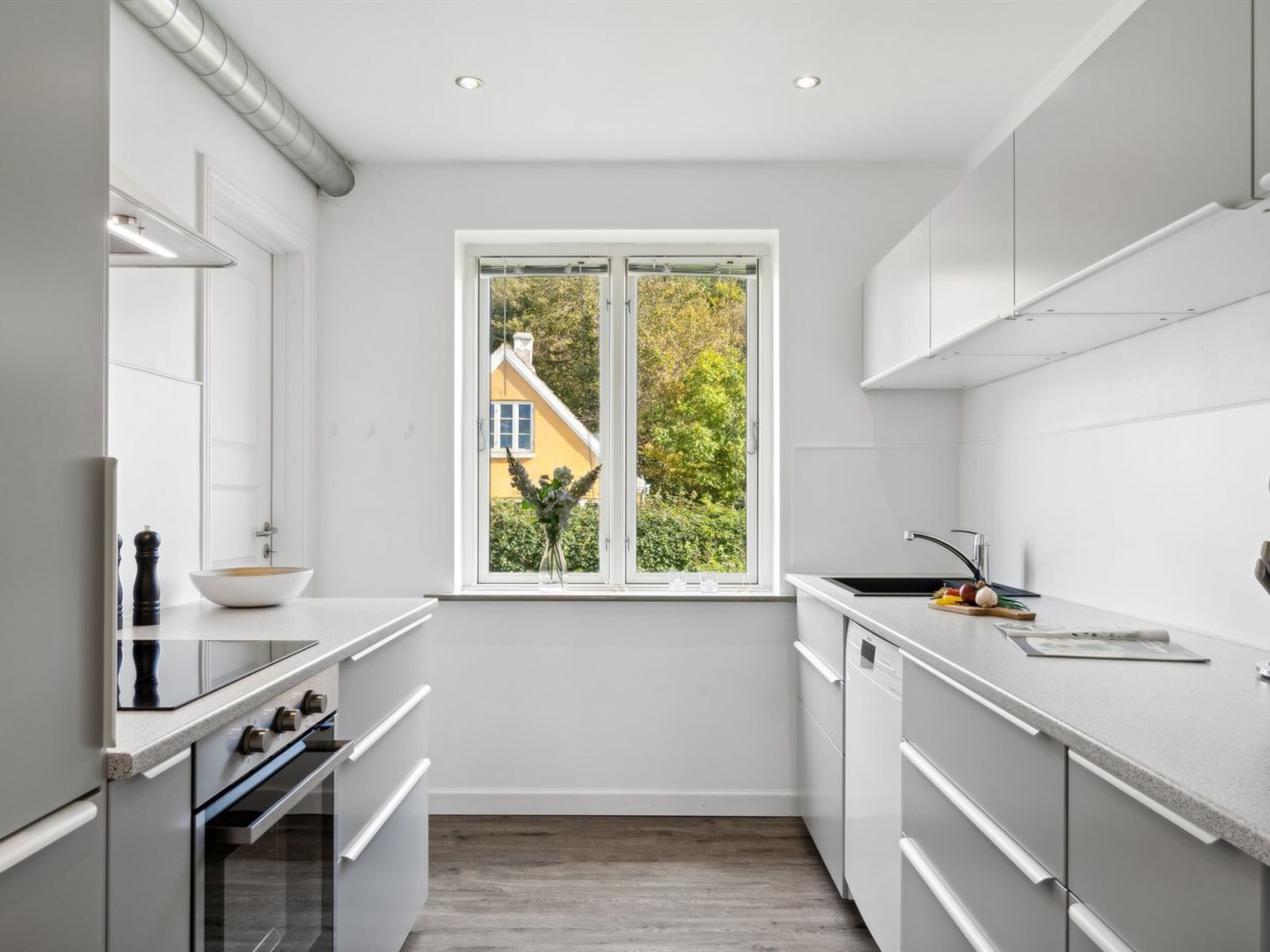 A modern kitchen with white cabinets, a double sink, and a window with a view of a yellow house and greenery.