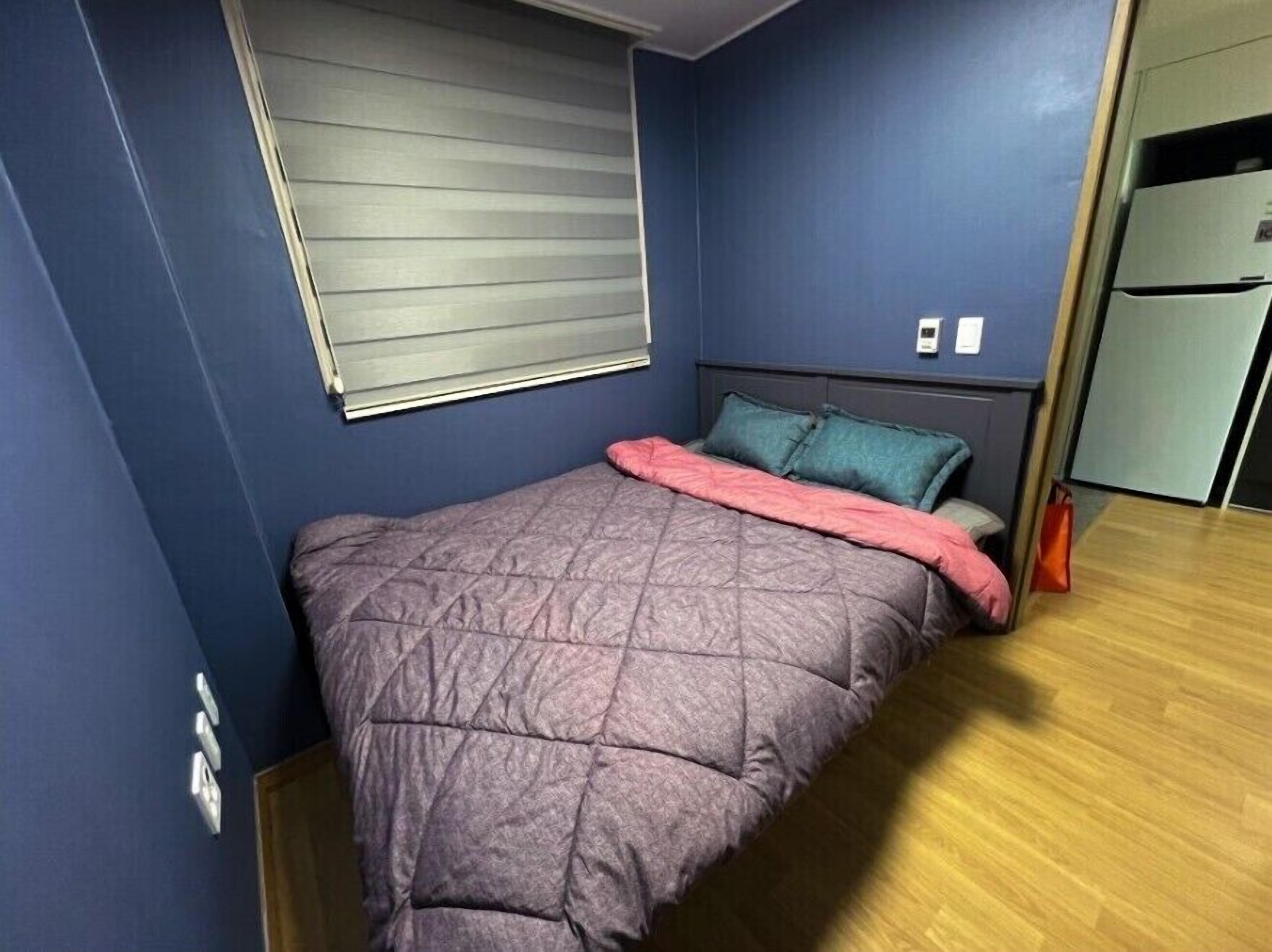 A bedroom with a bed, a window with blinds, a nightstand, and a small refrigerator.