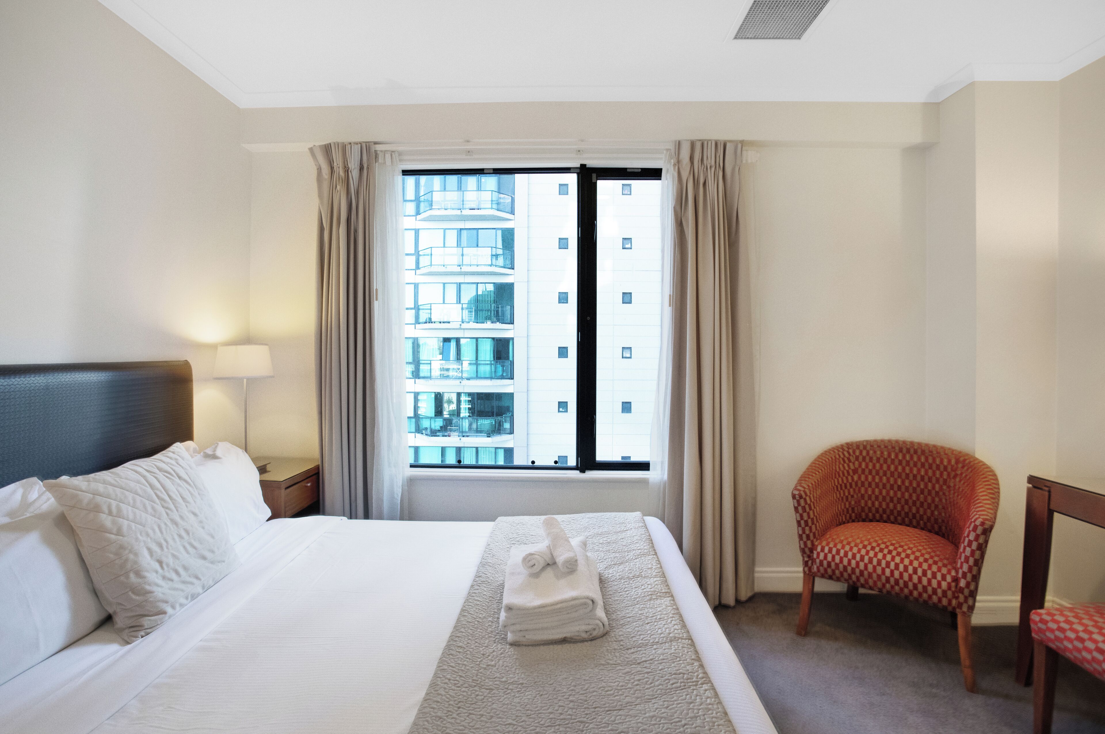 A hotel room with a bed, a chair, a window with curtains, and a view of a building outside.