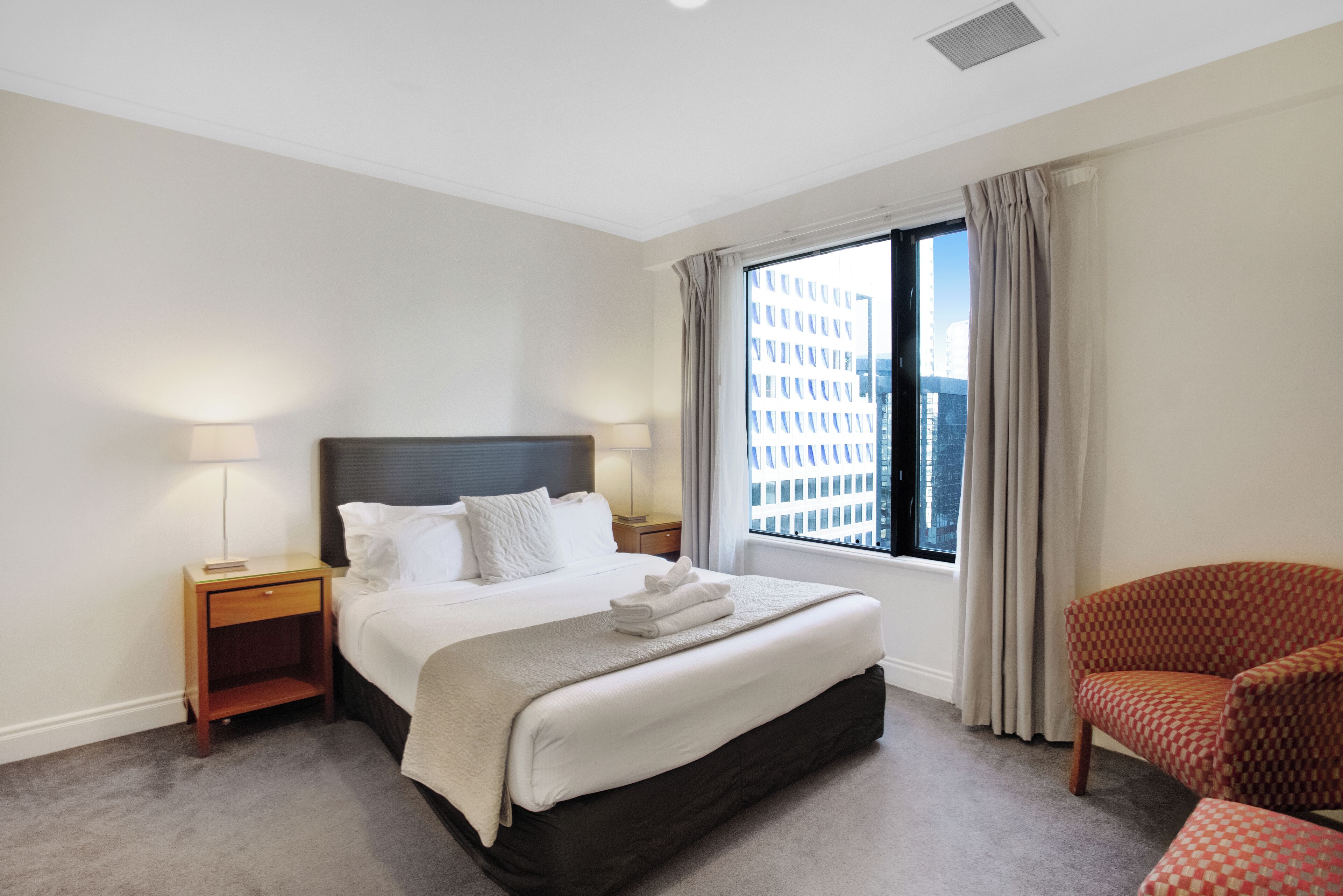 A hotel room with a large bed, a nightstand, a chair, and a window with a city view.