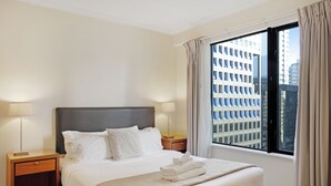 Una habitación de hotel con una cama grande, dos mesitas de noche, un ventanal con vistas a edificios y una cortina.