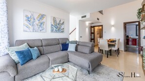 A modern living room with a grey sectional sofa, a glass coffee table, and two wall-mounted artworks. A dining area is visible in the background with a kitchen visible through an open door. - Luxury townhouse with rooftop Sauna & Jacuzzi REF062 (Fuengirola)