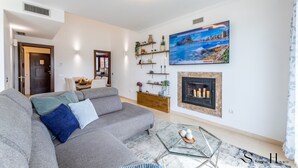 A modern living room with a grey sofa, a glass coffee table, and a fireplace. There is a painting on the wall and a wooden console table with decorative items. - Luxury townhouse with rooftop Sauna & Jacuzzi REF062 (Fuengirola)