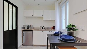 Cocina compacta con muebles blancos, horno empotrado y lavamanos. Hay un refrigerador pequeño, una mesa de comedor con platos azules y una planta en maceta.