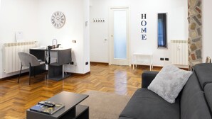 Moderni olohuone, jossa on musta sohva, sohvapöytä ja ruokailutila, jossa on tuoli ja pöytä. Seinällä on näkyvillä kello ja kyltti, jossa lukee ”KOTI”.