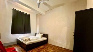 A small, neatly arranged bedroom with a single bed, a ceiling fan, a window with curtains, and a red chair. - Hotel B&B Heights (Guwahati)
