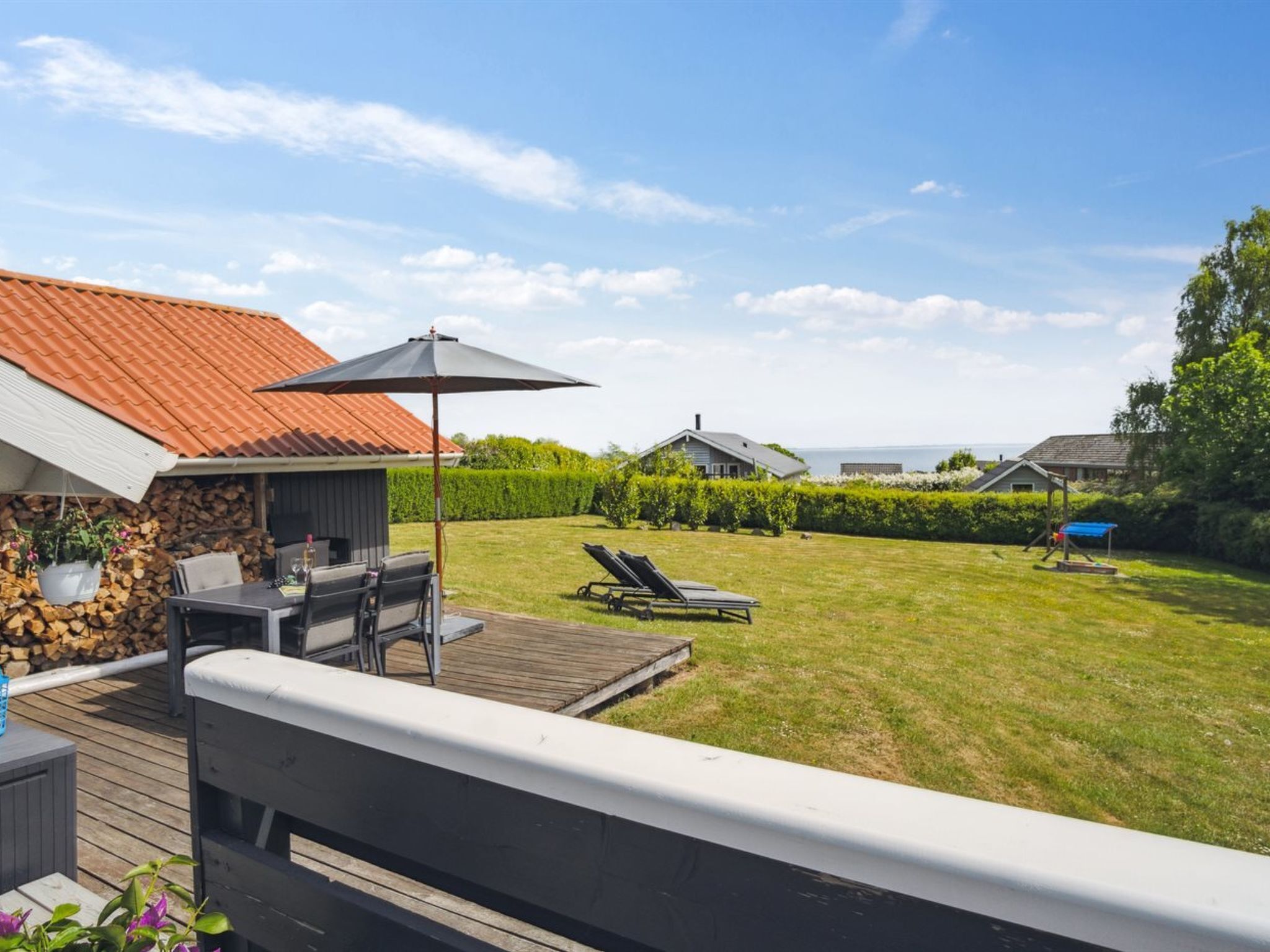 Una terraza con una parrilla y asientos exteriores, vista al patio trasero con césped y una zona de juegos, y una casa con techo rojo.