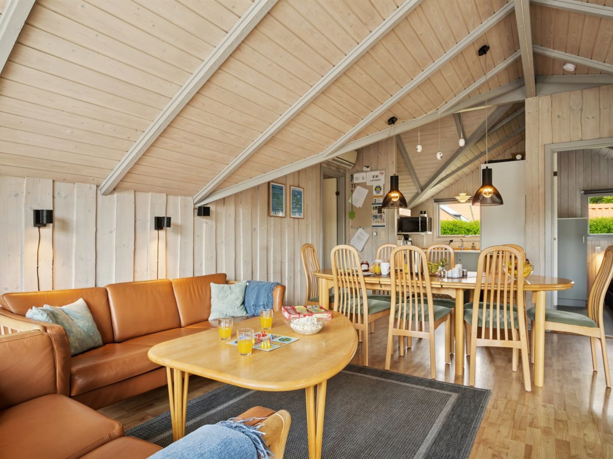 A cozy loft-style living room with a wooden ceiling, a brown leather sofa, a wooden dining table with chairs, and a kitchen area visible through an open door.