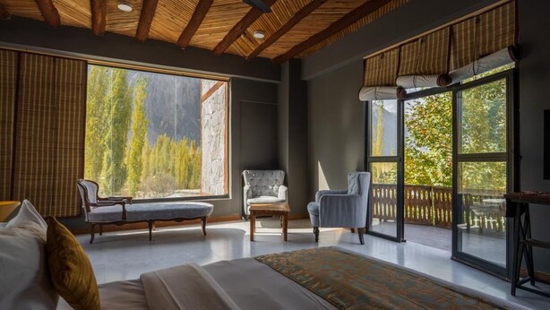 A spacious bedroom with a large bed, two chairs, and a view of trees through large windows. - PC Legacy Skardu (Skardu)