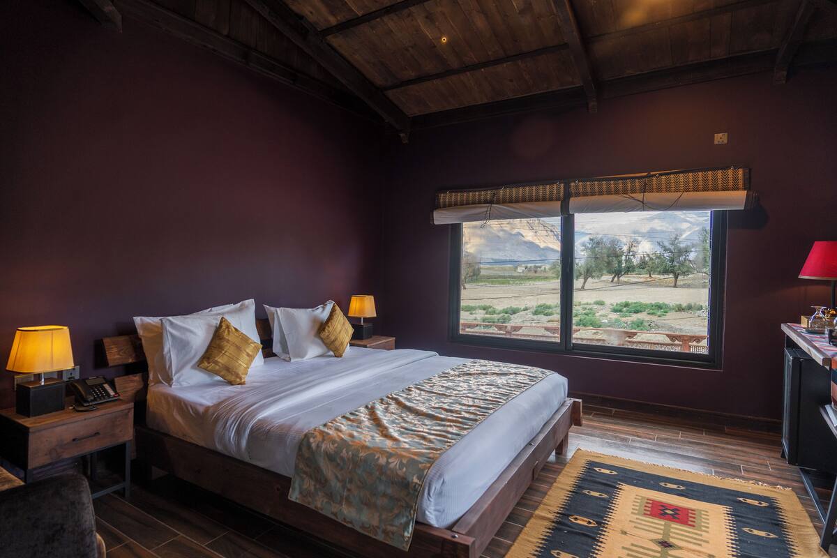 A bedroom with a large bed, wooden headboard, two bedside tables with lamps, a window with a view of the outdoors, and a red lamp on a desk.