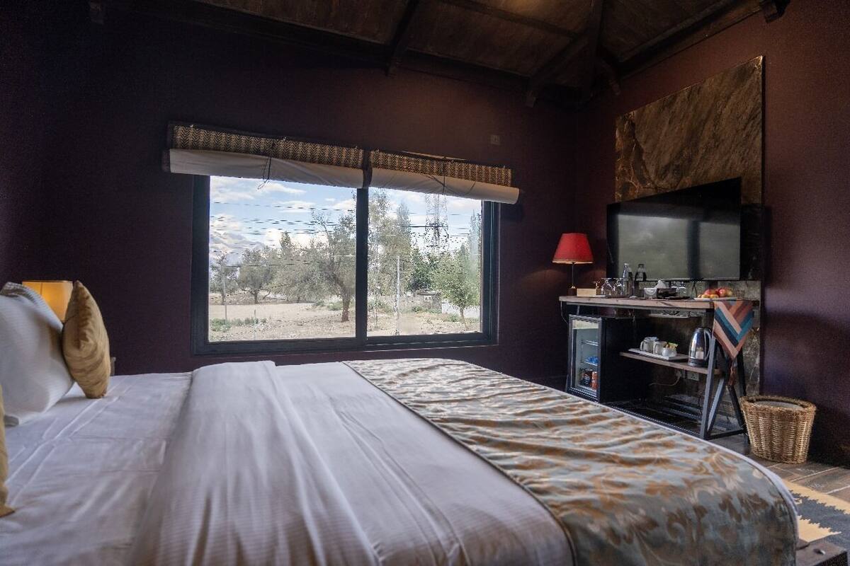 A bedroom with a bed, a window with a view of trees and mountains, a desk with a television, and a red lamp.