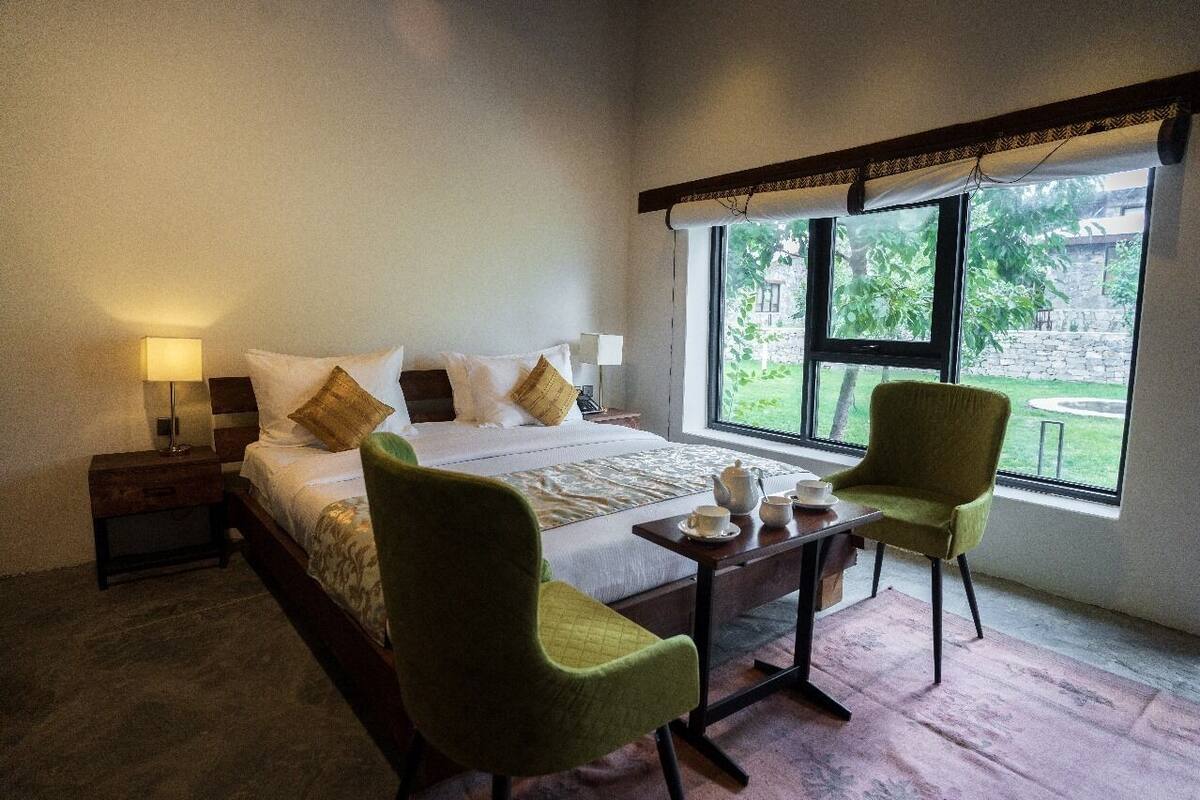 A bedroom with a bed, two chairs, a small table with cups and a teapot, and a window with a view of greenery.
