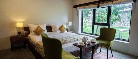 A bedroom with a bed, two chairs, a small table with cups and a teapot, and a window with a view of greenery.