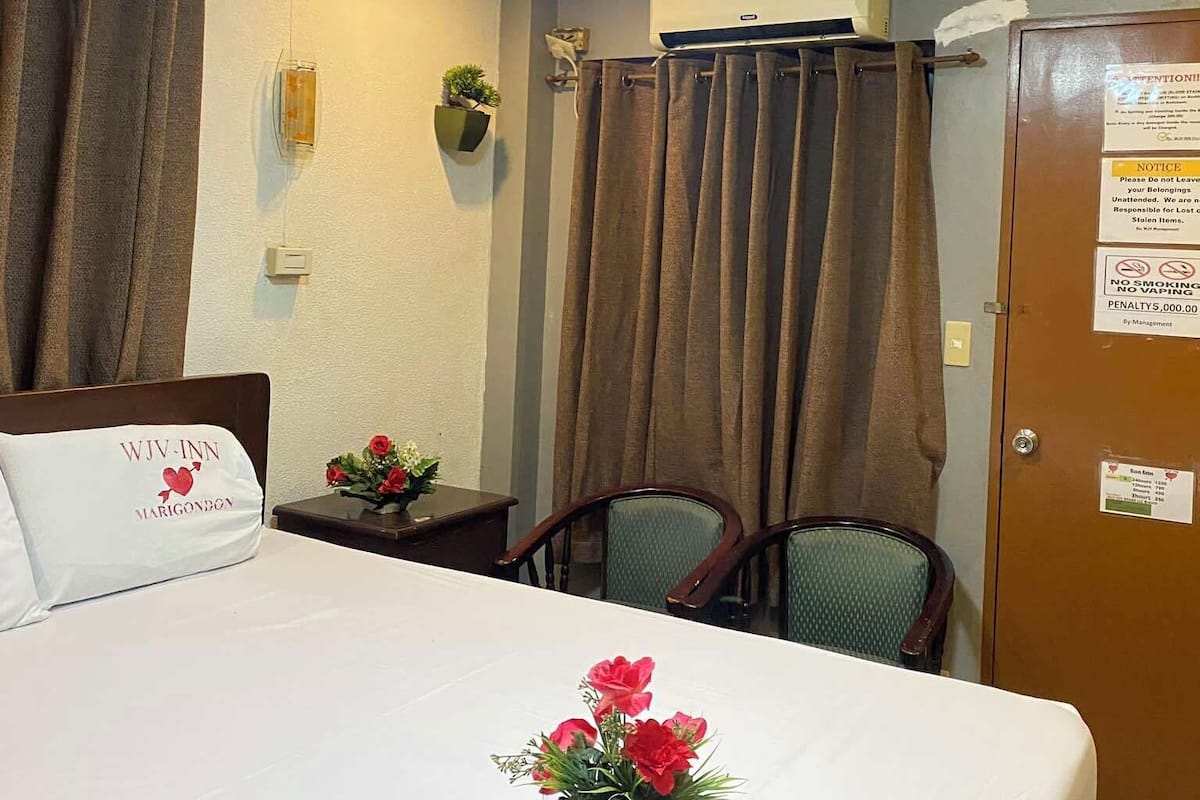 Ein Hotelzimmer mit Bett, Schreibtisch, Stuhl, einem kleinen Tisch mit Blumenvase, einer Wand-Klimaanlage und einer Tür mit einem Hinweis.