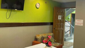 Ein Hotelzimmer mit grüner Wand, einer Uhr, einem an der Wand befestigten Fernseher, einer Tür mit einem Schild und einem Tisch mit einer Blumenvase.