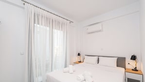 Habitación de hotel con cama, dos mesitas de noche, un ventanal con cortinas translúcidas y un equipo de aire acondicionado.