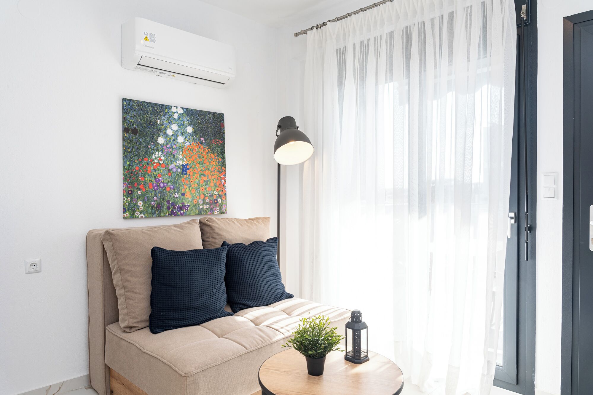 Un salon moderne avec un canapé beige, un tableau floral aux couleurs vives, une lampe sur pied, une petite table ornée d’une plante et d’une lanterne, et une porte-fenêtre coulissante.