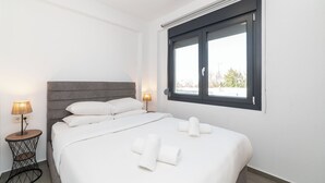 Ein ordentlich bezogenes Bett mit weißen Leinen und Handtüchern, ein grauer möbelbezogener Kopfteil und ein Fenster mit Blick auf Gebäude und Bäume.