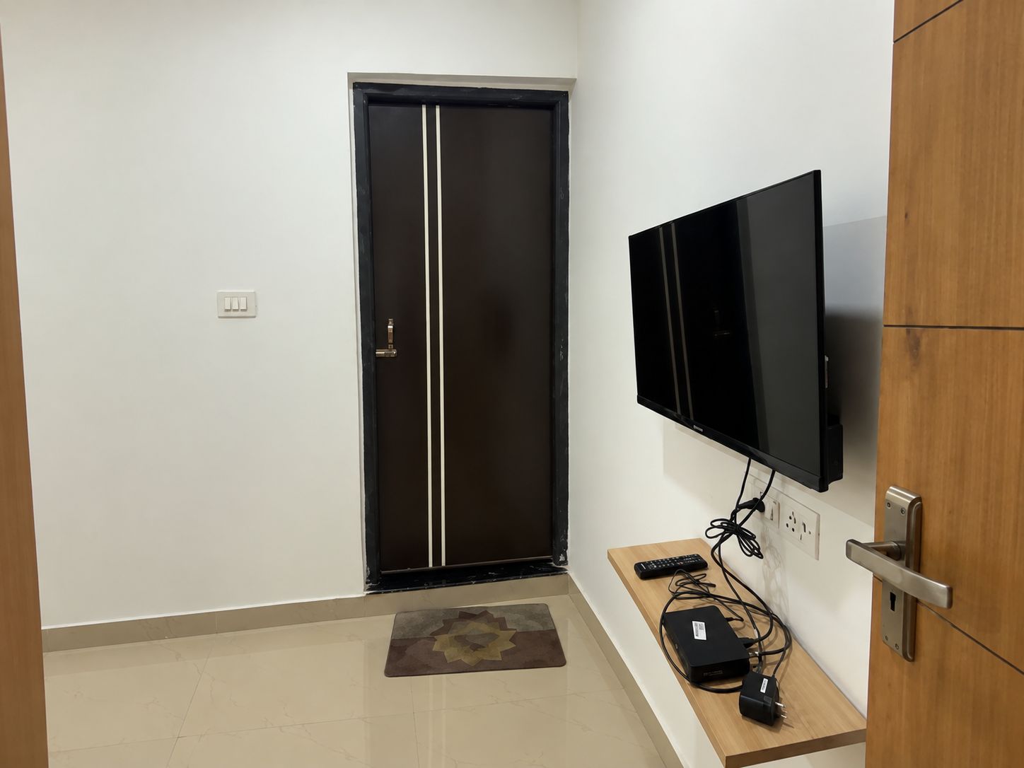 A room with a closed door, a mounted flat-screen TV, a wooden console table with a remote, and a power strip.