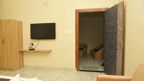 A room with a television mounted on the wall, a wooden wardrobe, a small wooden shelf, a cushioned chair, a marble floor, and a door leading to another room. - Hotel Royal Awas (Biswanath)