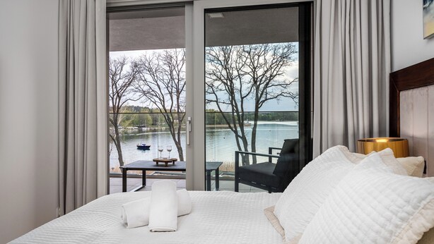 Une chambre d’hôtel dotée d’une grande fenêtre donnant sur un lac et des bateaux, un lit avec une literie blanche, une petite table avec deux verres à vin et une chaise.