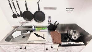 Una cocina con un lavamanos de acero inoxidable, una tabla de cortar, un cuchillo, una sartén, una espátula y una olla sobre una cocina de inducción.