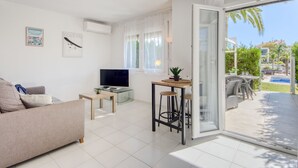 A modern living room with a sofa, a TV, a small table with a plant, and a glass door leading to an outdoor patio with a pool. - Puerto Rico by Interhome (Empuriabrava)