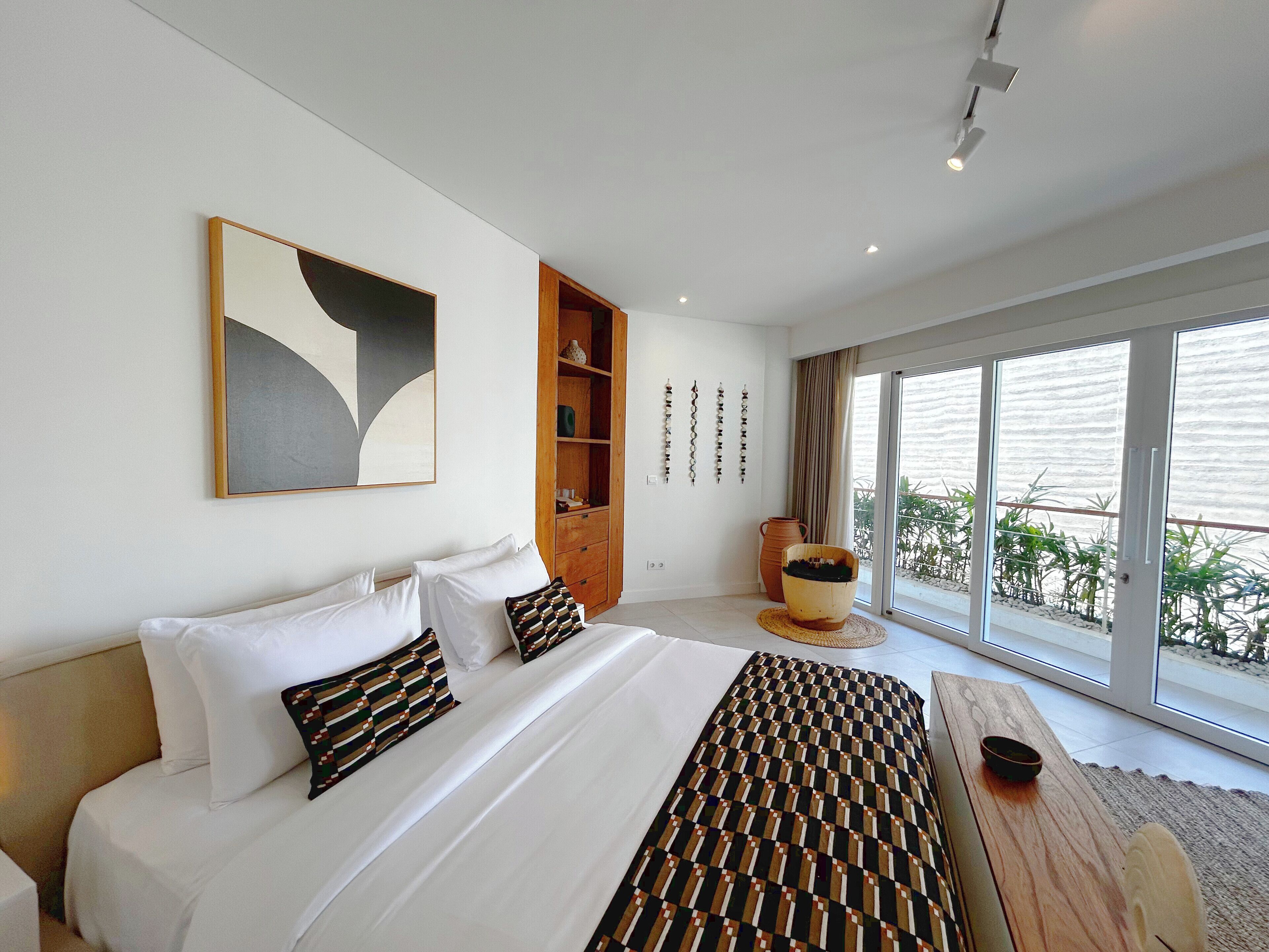 A modern bedroom with a large bed, a wooden cabinet, and a view of greenery through the window.