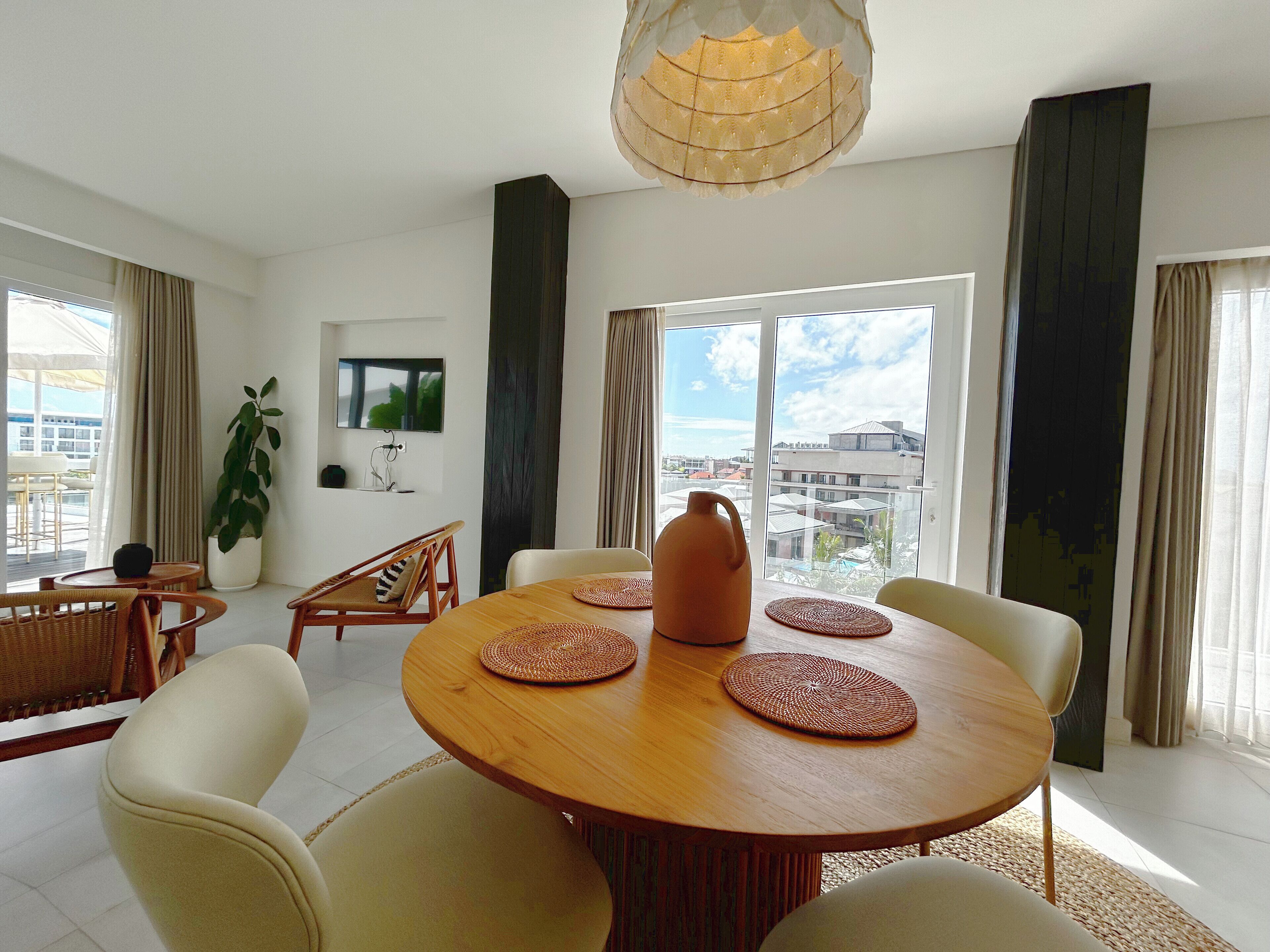 A modern dining area with a round wooden table, four chairs, and a large window offering a city view.